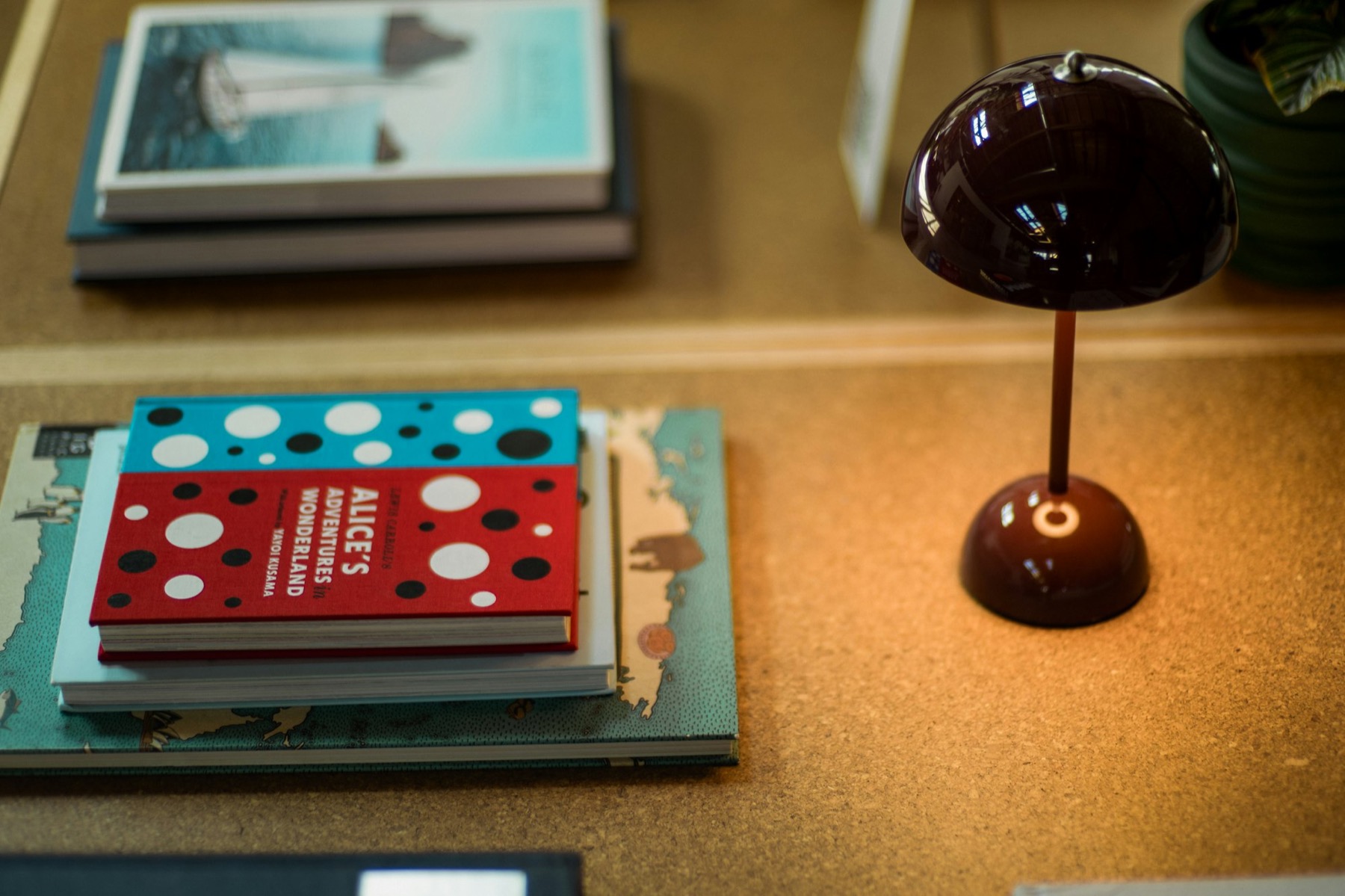 A stack of illustrated hardcover books beside a small plum-colored lamp on a cork desk