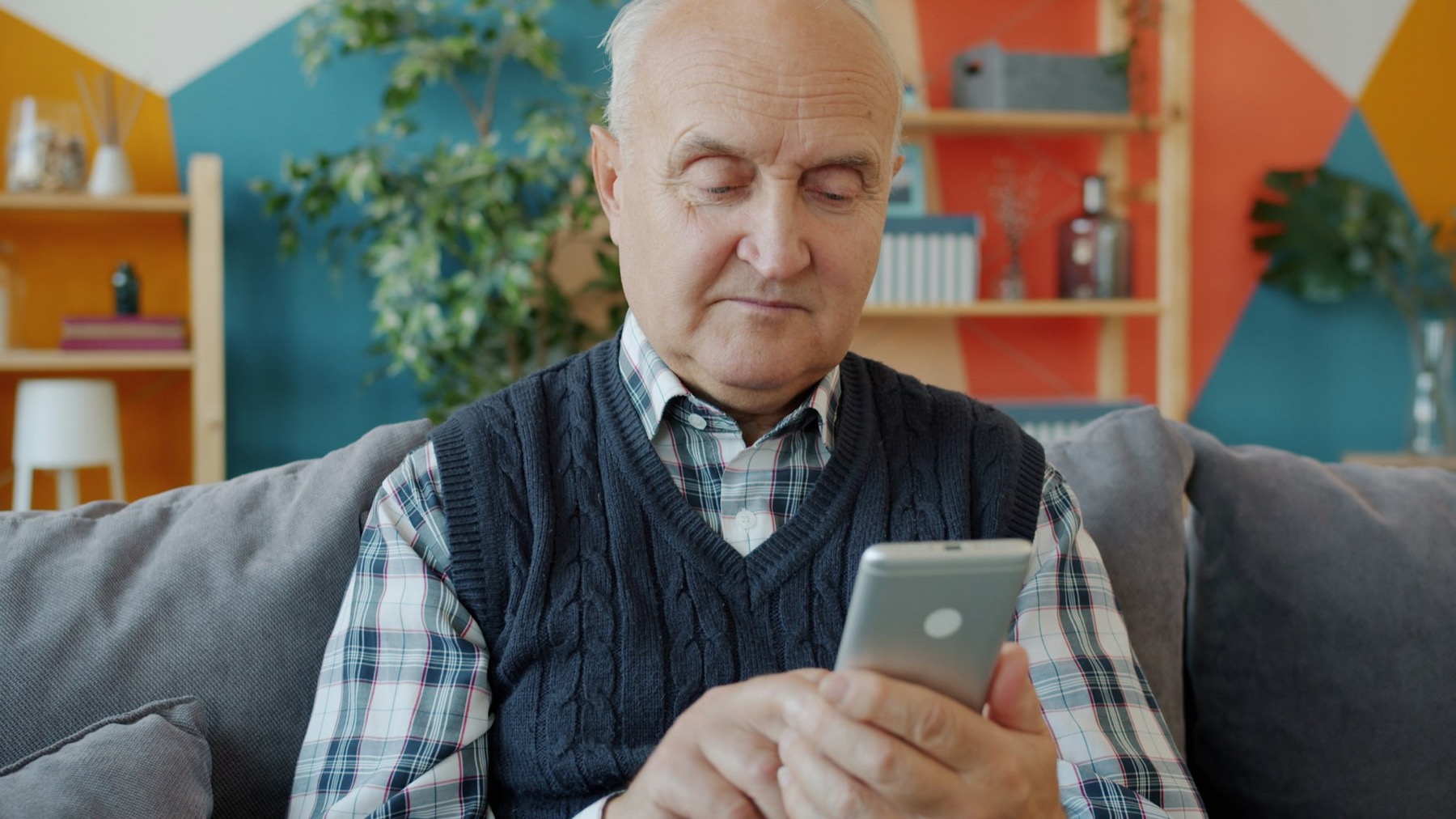 An older man on a sofa looking down at a smartphone in a bright living room