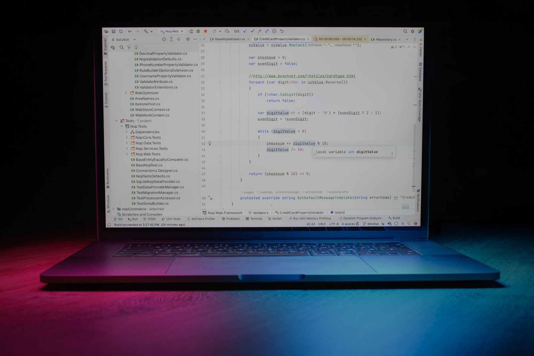 A laptop on a dark desk, its screen showing an open code editor, lit with pink and blue light.