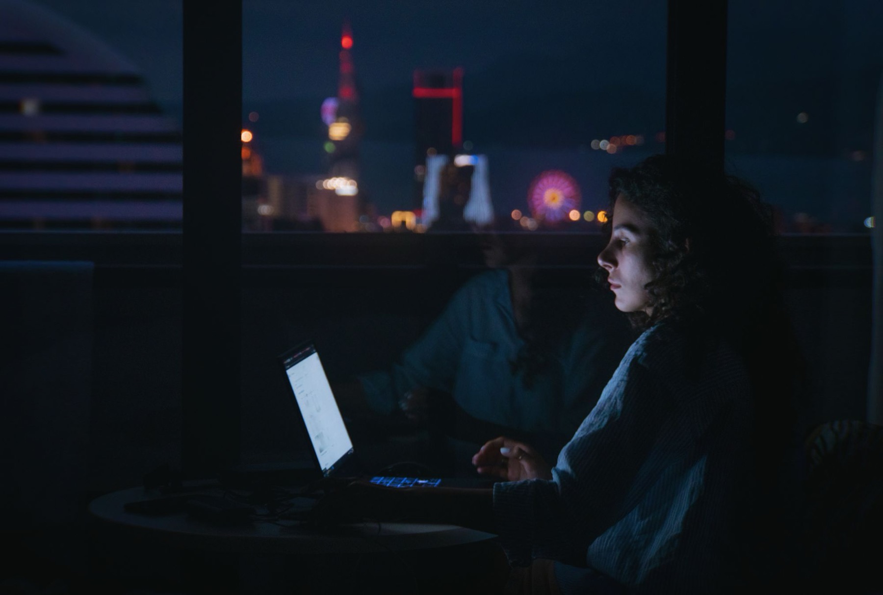 A woman works on a laptop in a dark room, lit by the screen, with a city skyline and lights through the window behind her.