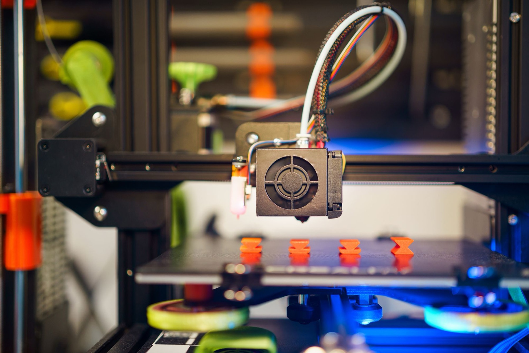 Close-up of a desktop 3D printer with its print head extruding orange plastic parts on a dark build plate, lit with blue and green accents.