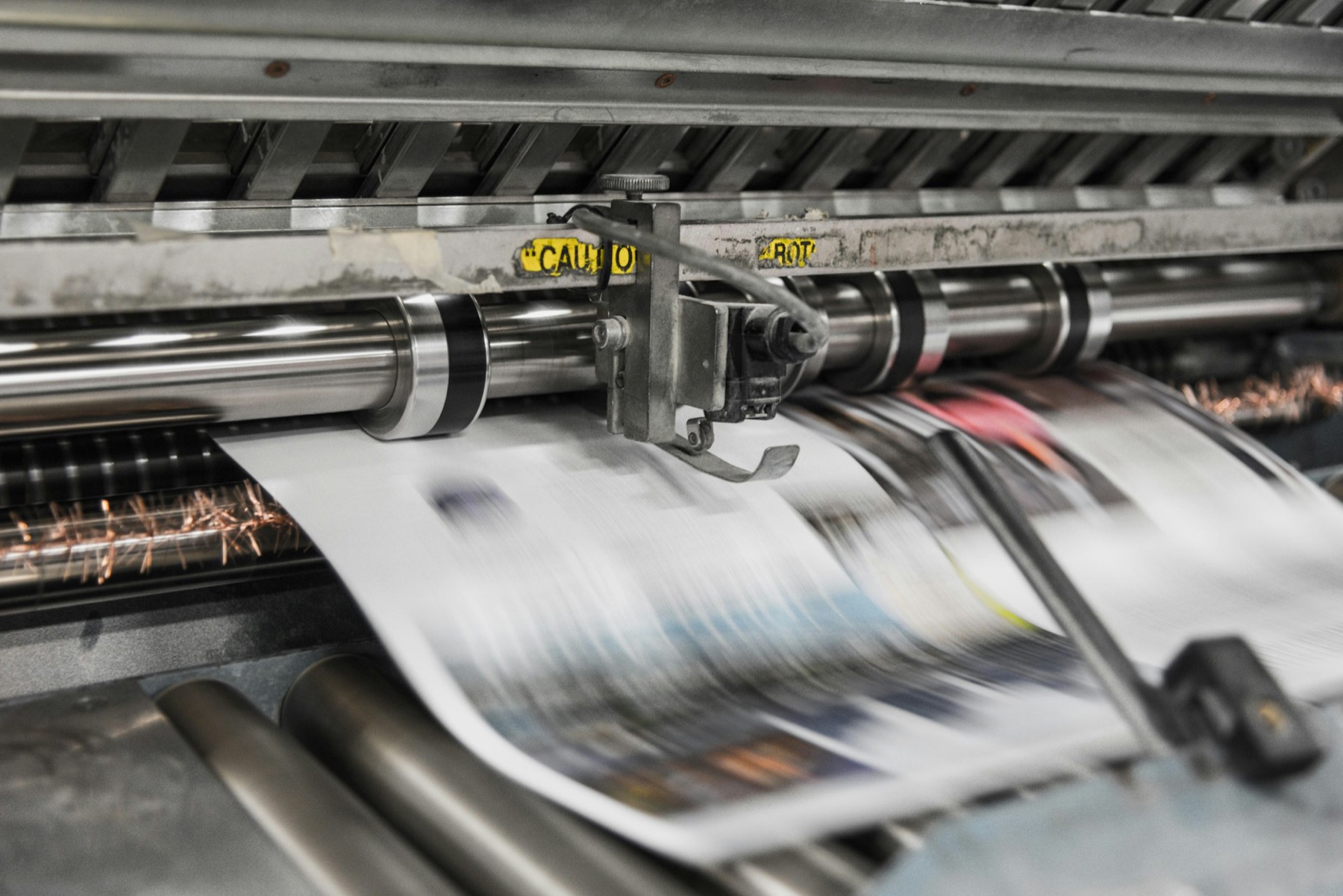 A printing press runs a newspaper through its rollers, the paper blurred with motion.